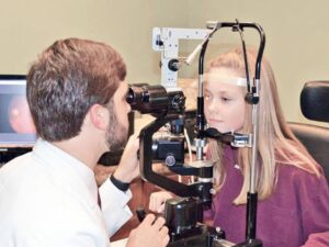 A young child sitting in an optometrist's chair having their eyes gently examined by a healthcare professional.