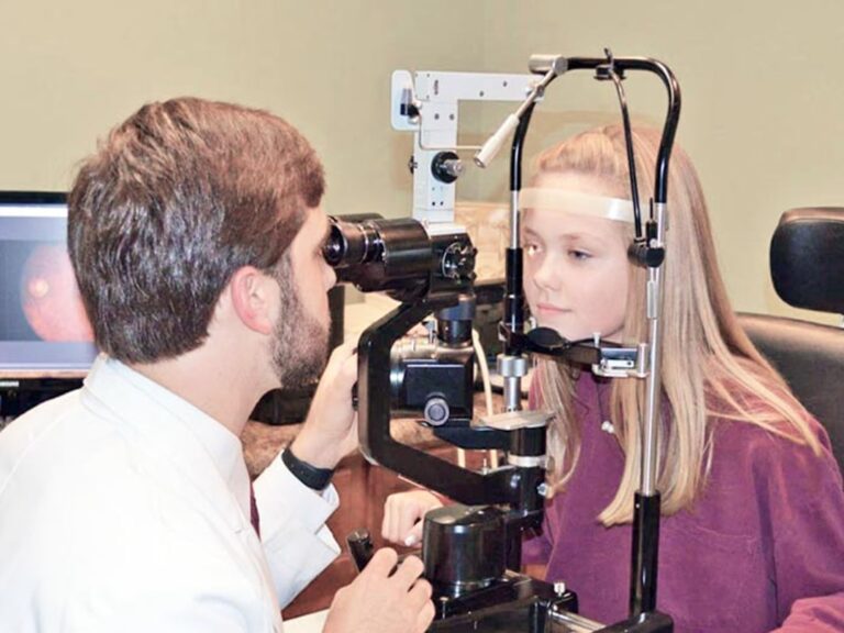 A young child sitting in an optometrist's chair having their eyes gently examined by a healthcare professional.